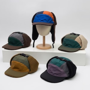 May include: Five multi-coloured trapper hats with ear flaps and flat brims. Each hat features a unique combination of fabric panels in shades of blue, orange, green, grey, brown, and black, paired with textured fleece or faux-shearling ear flaps.