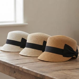 May include: Three straw hats arranged in a row, featuring a rounded crown and a short brim. Each hat is adorned with a wide black ribbon band tied into a bow on the side. The hats are colored in shades of cream, light tan, and medium brown.