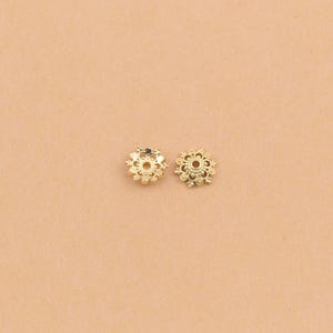 Può includere: Due piccole coppette per perline in metallo dorato di forma circolare, caratterizzate da un bordo smerlato delicato e un foro centrale. Questi componenti presentano un motivo testurizzato simile a un fiore che si irradia dal centro.