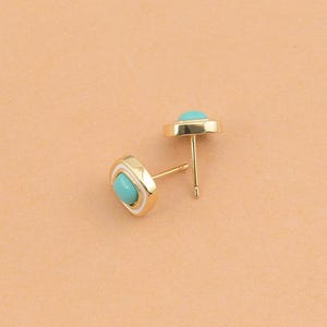 May include: A pair of gold stud earrings featuring a square-shaped frame with rounded corners. Each earring contains a central turquoise-colored stone surrounded by a thin white border, set within a polished gold-tone metal base with a post back.