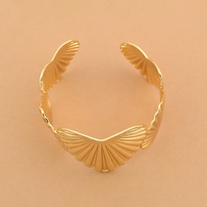 Può includere: Un bracciale aperto color oro caratterizzato da un motivo ripetitivo di elementi a forma di ventaglio. La fascia metallica è composta da segmenti radianti collegati che creano un bordo smerlato, formando una struttura circolare con apertura.