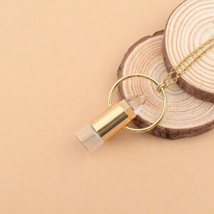 Puede incluir: Un collar de tono dorado con un colgante que presenta un cristal transparente y puntiagudo encerrado en un cilindro de metal dorado. El colgante cuelga de una fina cadena dorada que pasa a través de un anillo circular dorado en la parte superior.