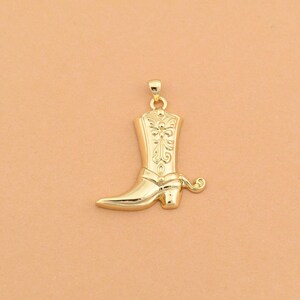 May include: A gold-toned metal pendant shaped like a cowboy boot. The boot features an intricate embossed floral or scrollwork pattern on the shaft and a small loop at the top for attachment to a chain or cord, with a polished, reflective finish.