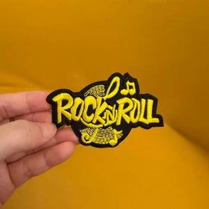 Peut inclure: Un écusson brodé présentant le texte ROCK N ROLL en fil jaune vif sur un fond noir. Le motif comprend des notes de musique stylisées et un élément graphique circulaire derrière le lettrage, créant un aspect audacieux et contrasté.