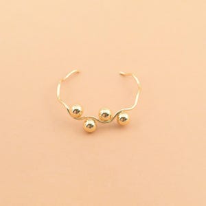 May include: A gold-toned open cuff bracelet featuring a thin, wavy wire band adorned with three smooth, polished spherical beads. The design is minimalist and symmetrical, showcasing a continuous metallic finish throughout the entire piece.