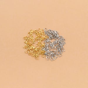 Puede incluir: Una colección de pequeñas piezas metálicas para joyería, específicamente terminales para cordones, dispuestas en un grupo circular. El surtido incluye piezas de tono dorado y plateado, cada una con un diseño en forma de U para asegurar cordones.