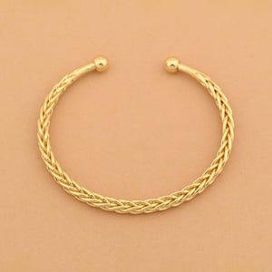 May include: A gold-toned cuff bracelet featuring a braided, rope-like texture throughout the band. The bracelet has an open design with a small, smooth spherical bead at each of the two terminal ends, creating a continuous circular shape.