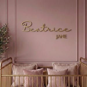 May include: A gold-colored wall sign featuring the name Beatrice in a cursive script font with the name Jane in a block font underneath. The sign is mounted on a dusty rose wall above a gold metal crib with matching pillows.