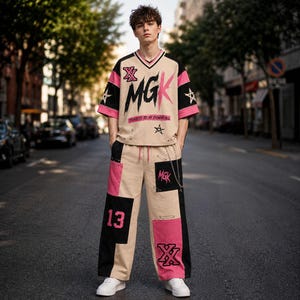 Könnte beinhalten: Ein beiges V-Ausschnitt-T-Shirt mit rosa und schwarzen Akzenten, das den Text MGK und Tickets to My Downfall zeigt, kombiniert mit einer passenden beigen, rosa und schwarzen Color-Block-Hose mit der Nummer 13, Sternen und einer silbernen Kette.