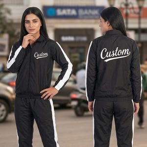 May include: Black tracksuit with white stripes on the sleeves and legs. The jacket has a zipper closure and the word "Custom" in white script on the chest and back. The model is wearing the tracksuit.