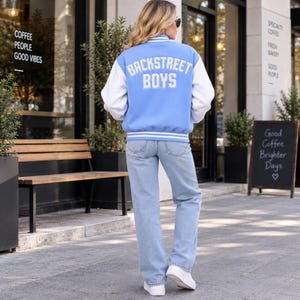 Könnte beinhalten: Eine hellblaue und weiße Collegejacke mit dem Schriftzug BACKSTREET BOYS in weißen Buchstaben auf dem Rücken, kombiniert mit einer hellblauen Jeans und weißen Turnschuhen. Die Jacke hat weiße Ärmel sowie einen gestreiften Rippkragen und Saum.