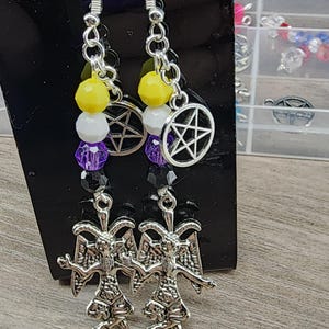 Puede incluir: Pendientes colgantes con dijes de Baphomet en tono plateado, dijes circulares de pentagrama y cuentas facetadas en amarillo, blanco, morado y negro. Los pendientes cuelgan de ganchos de metal plateado y se exhiben en una tarjeta negra.