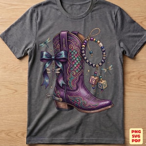 May include: A charcoal gray t-shirt featuring a graphic of a purple cowboy boot with intricate teal and pink scale patterns. The boot is adorned with a large blue bow, a dragonfly, and a beaded charm bracelet with blocks spelling ENCHANT.