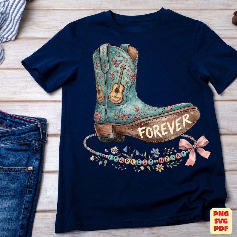 May include: A navy blue short-sleeve t-shirt featuring a graphic of a teal cowboy boot decorated with flowers, butterflies, and a guitar. The word FOREVER is printed on the boot, and a string of beads spells FEARLESS HEART below it.