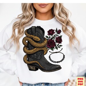 May include: A white sweatshirt featuring a graphic print of a black cowboy boot wrapped in a golden snake, accompanied by dark red roses and a beaded bracelet with the text "KARMA IS REAL" printed in black letters on the beads.
