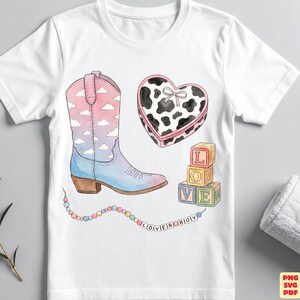May include: A white t-shirt featuring a watercolor-style graphic of a cowboy boot with a pink and blue cloud pattern, a heart-shaped box with a black and white cow print, three stacked alphabet blocks, and a string of beads spelling LOVER BOY.