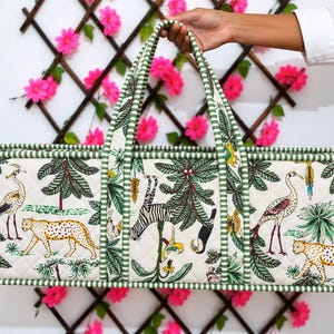 May include: A rectangular quilted tote bag featuring a cream-colored fabric with a jungle-themed print of leopards, cranes, and tropical trees. The bag has green and white striped fabric trim and two matching striped handles for carrying.