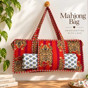 May include: A rectangular fabric bag featuring a vibrant red and orange striped pattern with tiger illustrations. The bag has two long handles and contains two rolled white cloths with blue floral prints. Text on the image reads Mahjong Bag.
