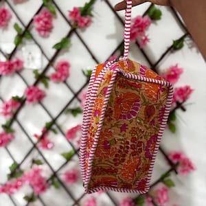 Könnte beinhalten: Eine rechteckige Stofftasche mit beigefarbener Basis und einem lebendigen Blumenmuster in Orange- und Rosatönen. Die Tasche ist an den Kanten mit weiß-rosa gestreiften Paspeln versehen und verfügt über eine passende gestreifte Handschlaufe.