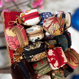 Peut inclure: Une collection de petites pochettes matelassées à fermeture éclair avec divers motifs de tissus colorés, incluant des fleurs, des imprimés animaliers et des textures unies. Chaque pochette possède un mousqueton doré sur une languette en cuir marron.