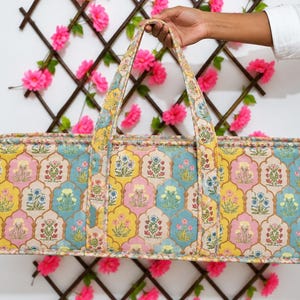 May include: A rectangular quilted duffel bag featuring a repeating pattern of floral motifs inside scalloped shapes. The fabric displays a color palette of yellow, light blue, and pink. The bag includes two long fabric handles for carrying.