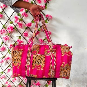 May include: A rectangular quilted duffel bag in bright pink featuring a repeating tiger print pattern. The bag has two long handles with a white and brown striped design and a zipper closure along the top edge. The fabric has a vertical stitched texture.
