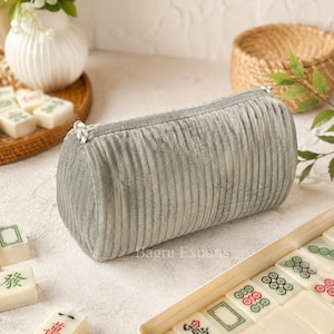 May include: A cylindrical pencil case made of grey, vertically ribbed velvet fabric with a silver zip closure. The case is positioned near a set of white mahjong tiles and a white vase containing white flowers.