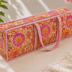 Puede incluir: Una bolsa de almacenamiento rectangular de tela acolchada con un estampado floral en tonos rosa y naranja sobre un fondo beige. La bolsa tiene un cierre de cremallera, ribetes a rayas rosas y blancas en los bordes y dos asas a juego.