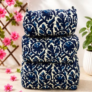 Puede incluir: Una pila de tres edredones plegados con un estampado floral en azul oscuro y crema. La tela tiene una textura acolchada visible con líneas de costura verticales que recorren la superficie de cada capa textil plegada.