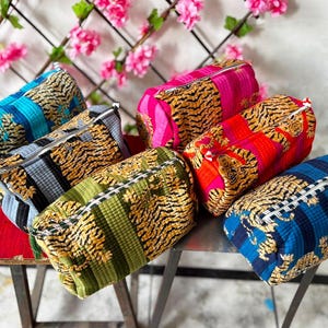 Könnte beinhalten: Eine Sammlung von sechs rechteckigen Reißverschlusstaschen mit einem sich wiederholenden Tigermuster auf strukturiertem Stoff. Die Taschen sind in verschiedenen Farben erhältlich, darunter Blau, Grau, Grün, Pink, Rot und Dunkelblau, jeweils mit silbernem Reißverschluss.