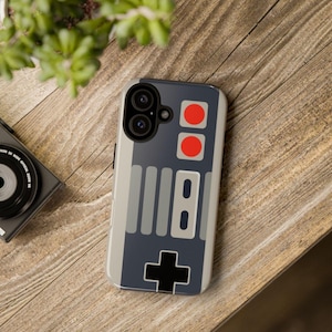 May include: A smartphone case featuring a retro video game controller design. The case has a gray background with red buttons, gray stripes, and a black directional pad. The phone case is on a wooden surface.