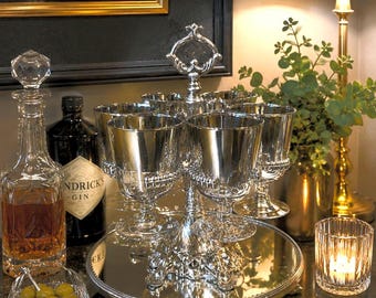 May include: A silver-plated serving tray with six stemmed glasses, a crystal decanter, and a small bowl of olives. A bottle of gin and a lit candle are also present. The arrangement is set on a dark countertop.