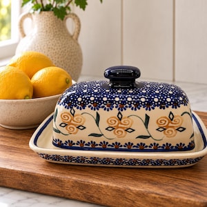 May include: A rectangular ceramic butter dish with a lid, featuring a cream-colored base decorated with hand-painted yellow floral swirls, dark green leaves, and a border of small blue flowers and white dots, topped with a dark blue handle.