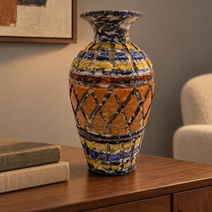 May include: A ceramic vase featuring a textured surface with a multi-colored pattern. The design includes horizontal bands of yellow and blue squares, a central section with an orange diamond grid, and a dark, mottled finish on the neck and base.