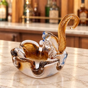 May include: A clear and amber-colored glass bowl featuring a sculpted squirrel figure attached to the rim. The bowl has a wavy, organic edge with swirling brown and white patterns throughout the glass, creating a decorative vessel for small items.