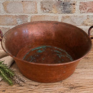 May include: A large, round, hammered copper basin featuring two riveted metal handles on opposite sides. The exterior has a warm, metallic copper finish, while the interior shows signs of oxidation with visible patches of green patina.