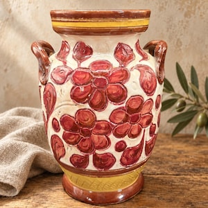 May include: A ceramic vase featuring a cream-colored body adorned with hand-painted red floral motifs and leaves. The vessel includes two side handles, a wide rim with yellow and brown bands, and a textured yellow base with a brown trim.