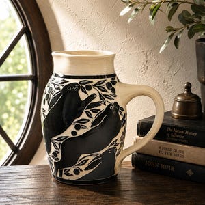 May include: A cream-colored ceramic pitcher featuring a black sgraffito design of three rabbits surrounded by leafy branches. The pitcher has a wide handle, a flared rim, and a smooth finish, showcasing a high-contrast black and white aesthetic.