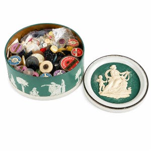 May include: A round green metal tin filled with various loose buttons and small containers, featuring white neoclassical relief figures on the exterior. The lid is detached, displaying a white relief of a seated figure and a child on a green background.