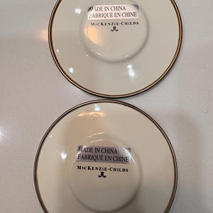 May include: Two off-white ceramic saucers with a gold rim. Each saucer has a central recessed area and a sticker that reads "MADE IN CHINA FABRIQU&Eacute; EN CHINE MACKENZIE-CHILDS".