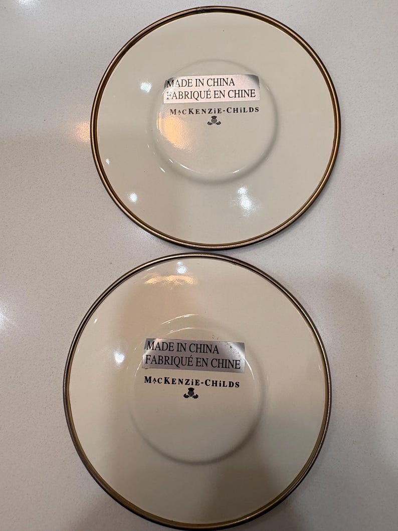 May include: Two off-white ceramic saucers with a gold rim. Each saucer has a central recessed area and a sticker that reads "MADE IN CHINA FABRIQU&Eacute; EN CHINE MACKENZIE-CHILDS".