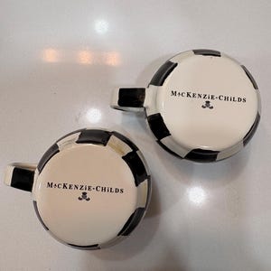 May include: Two ceramic mugs with black and white checkered patterns and black handles. The bottom of each mug features the text "MacKenzie-Childs" in black. The mugs are set against a white surface.