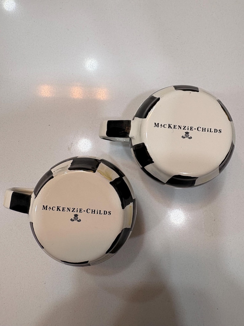 May include: Two ceramic mugs with black and white checkered patterns and black handles. The bottom of each mug features the text "MacKenzie-Childs" in black. The mugs are set against a white surface.