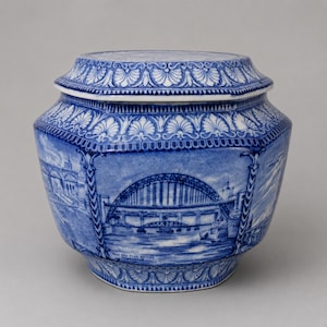 May include: A blue and white ceramic jar with a lid, featuring detailed illustrations of the Tyne Bridge and other architectural landmarks. The vessel has a hexagonal shape with decorative border patterns and text labels below the bridge imagery.