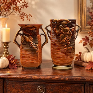 May include: Two brown ceramic vases with textured, horizontal ridged surfaces and ornate handles. Each vase features a raised, three-dimensional relief of grape leaves and clusters of grapes on the front, finished in a warm, autumnal tone.