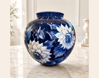 May include: A round ceramic vase with a deep cobalt blue glaze featuring hand-painted white floral motifs and leaves. The neck of the vase is accented with a thin gold-colored band and a delicate repeating pattern around the upper rim.