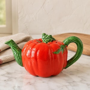 May include: A ceramic teapot shaped like a bright red pumpkin with a glossy finish. The teapot features a green textured handle, a green spout shaped like a leaf, and a green stem lid handle. The body has vertical ridges mimicking a pumpkin.