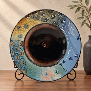 May include: A round ceramic decorative plate featuring a deep black center with a spiral design. The wide rim displays a gradient of yellow, blue, and green with textured patterns, swirling motifs, and small blue dots, supported by a metal stand.