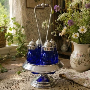 May include: A silver-toned metal condiment server featuring a central carrying handle and a circular base. The server holds four cobalt blue glass jars, each topped with a polished silver-toned metal lid and a small integrated spoon.