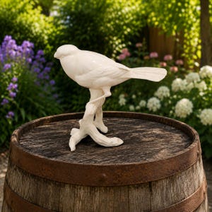 Może przedstawiać: Błyszcząca biała ceramiczna figurka ptaka siedząca na teksturowanej gałęzi. Ptak jest przedstawiony z profilu, z gładkimi, wyrzeźbionymi piórami i długim ogonem. Rzeźba została wykonana z jednego kawałka białego materiału.